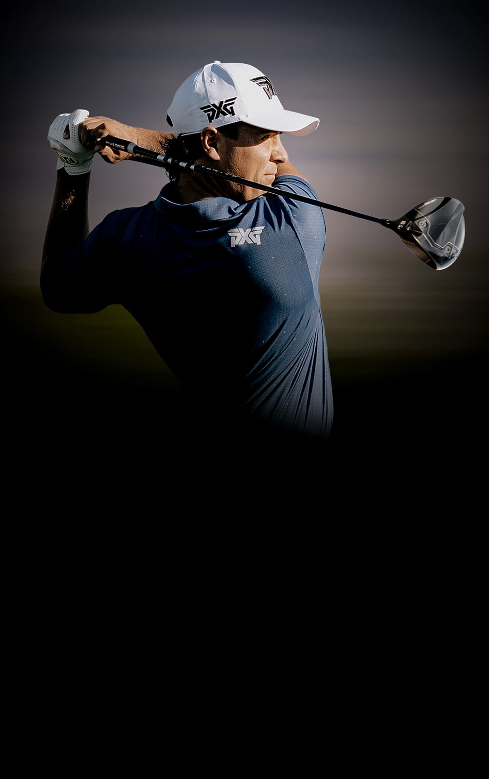 A golfer in a navy blue shirt and white cap swings a driver, captured in dramatic low-key lighting against a dark background.