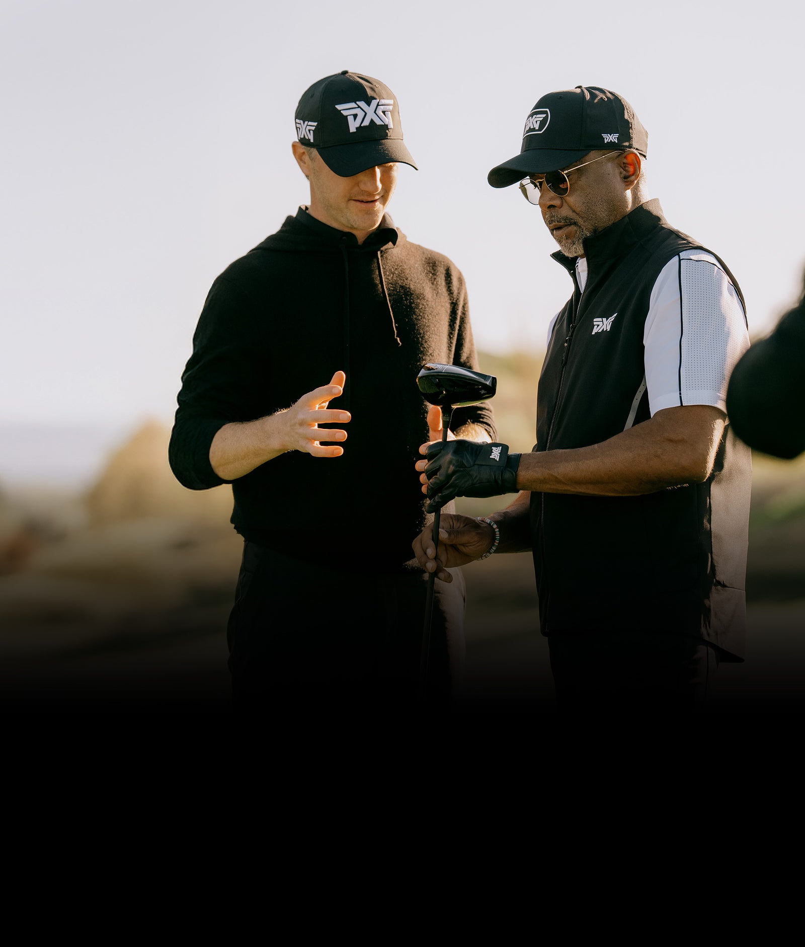 Two men in black jackets and hats with PXG logos, one holding a golf club, against a blurred outdoor background.