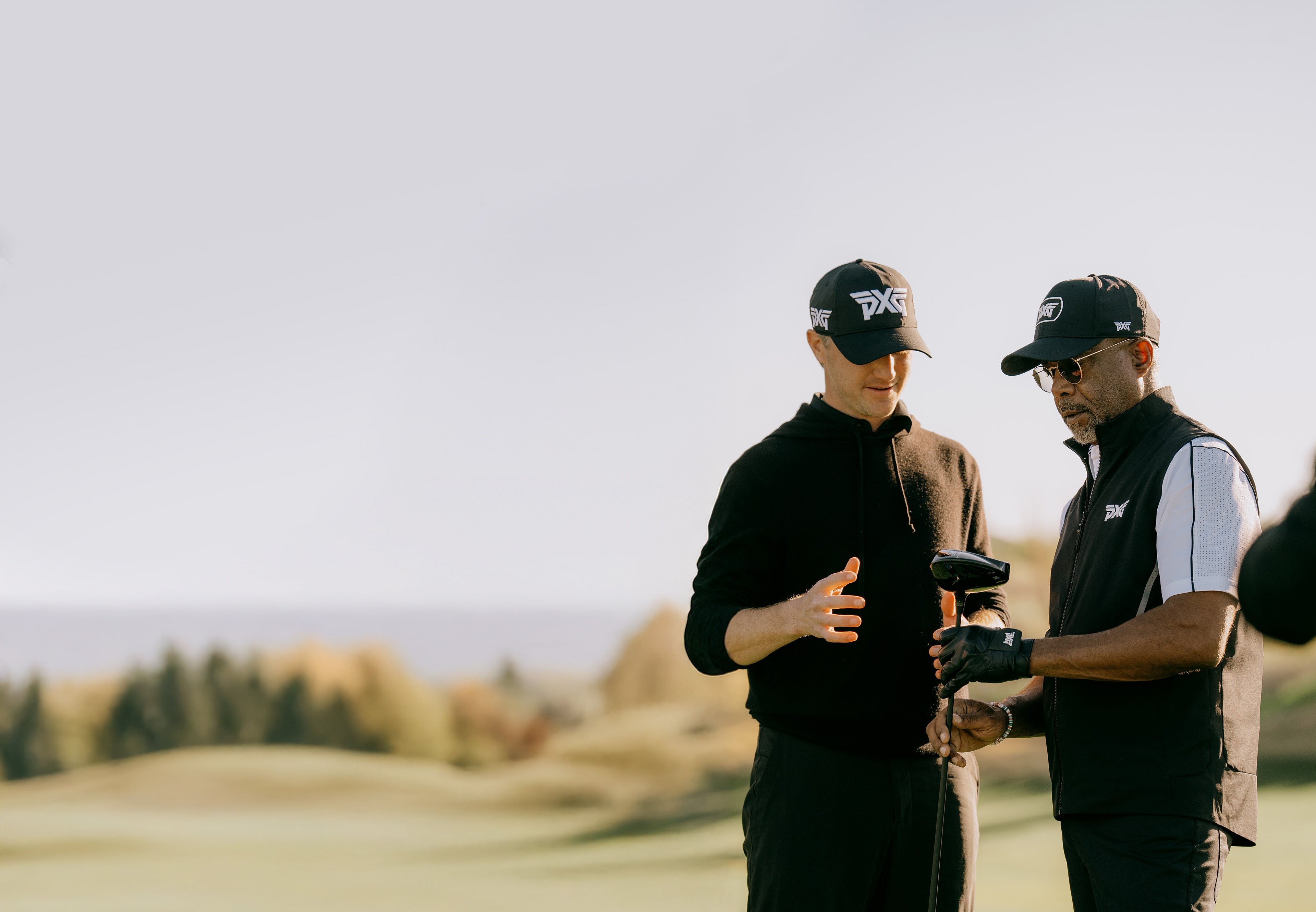Two men on a golf course discussing a golf club with a scenic background