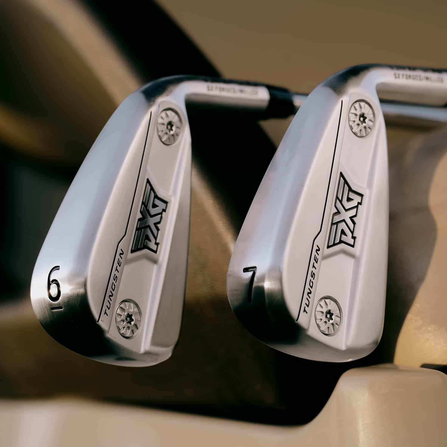 Close-up of two silver PXG golf irons, numbered 6 and 7, with black and white branding, resting in a beige and dark brown golf bag.