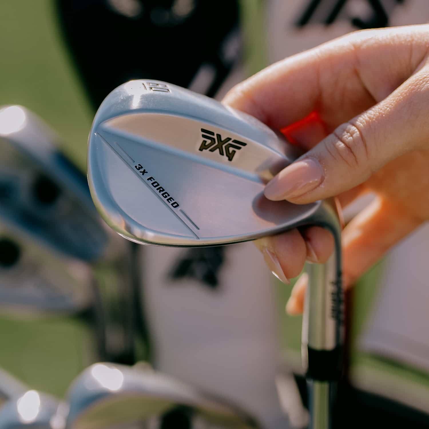 A close-up of a person's hand holding a PXG 3X Forged golf wedge, showing the silver and white club head with black branding, against a blurred outdoor background.
