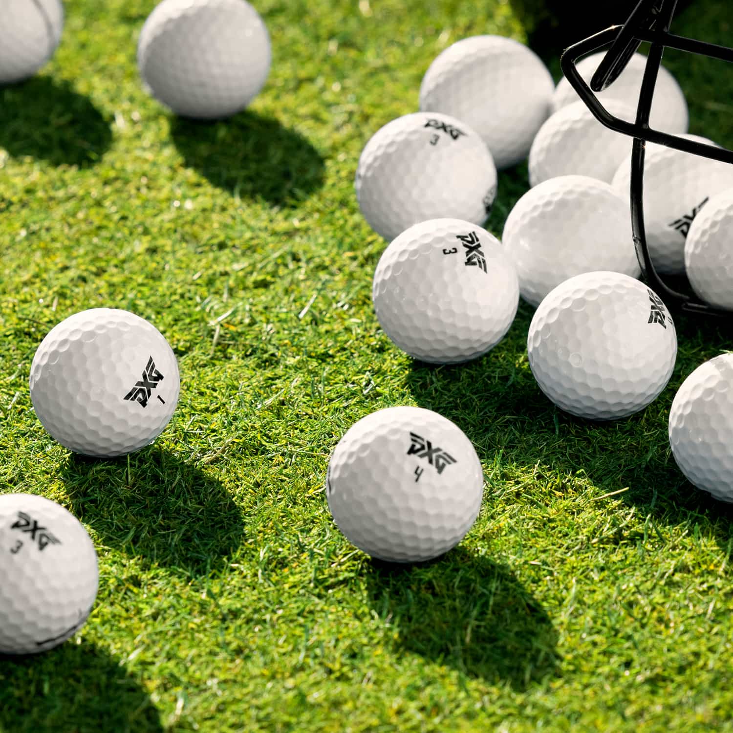 White golf balls with black printed logos scattered on bright green grass, with a black metal golf basket partially visible on the right.