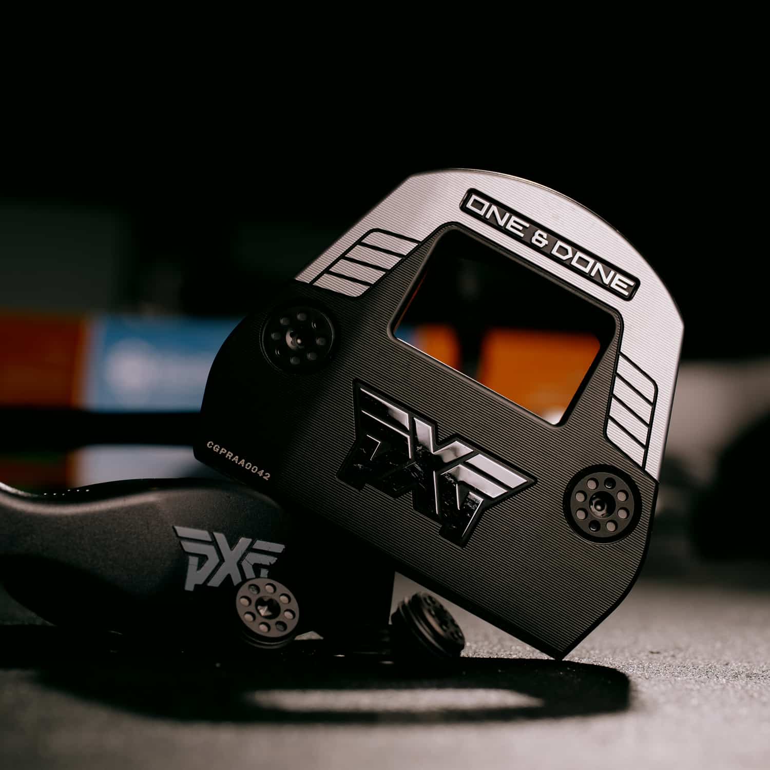 A close-up product shot of a PXG One & Done red dot sight with a black and white textured finish, resting on a dark surface with a blurred background.