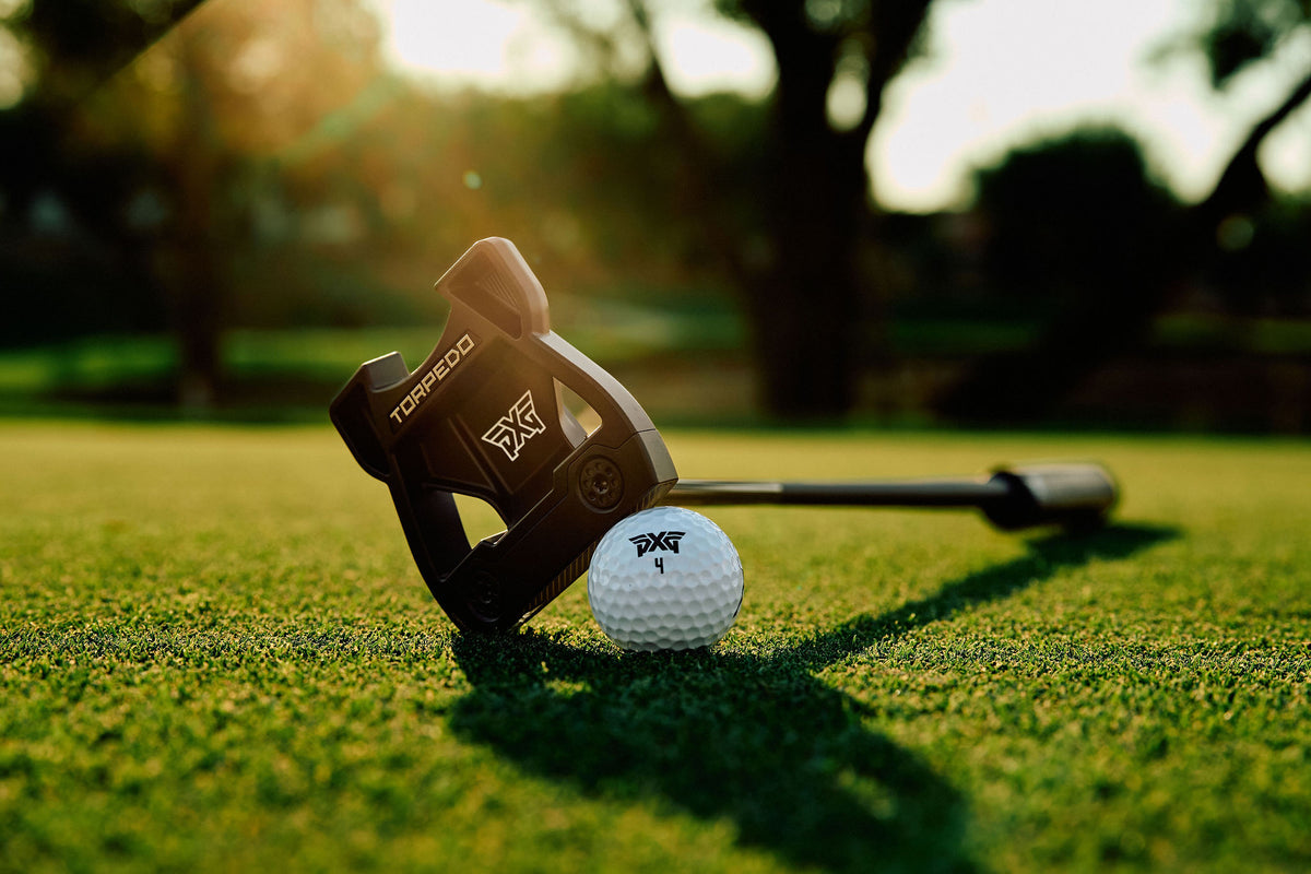 A Torqued golf putter head resting on a golf green next to a white golf ball, with a blurred outdoor golf course background and warm sunlight.