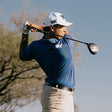 Golfer in blue shirt and white cap swinging a golf club with a blurred natural background